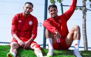 Hakim Ziyech et un coéquipier assis décontractés sur un talus herbeux, portant tous deux les tenues d'entraînement rouges du Wydad Casablanca, profitant d'une journée ensoleillée.