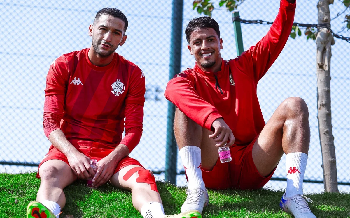 Hakim Ziyech et un coéquipier assis décontractés sur un talus herbeux, portant tous deux les tenues d'entraînement rouges du Wydad Casablanca, profitant d'une journée ensoleillée.