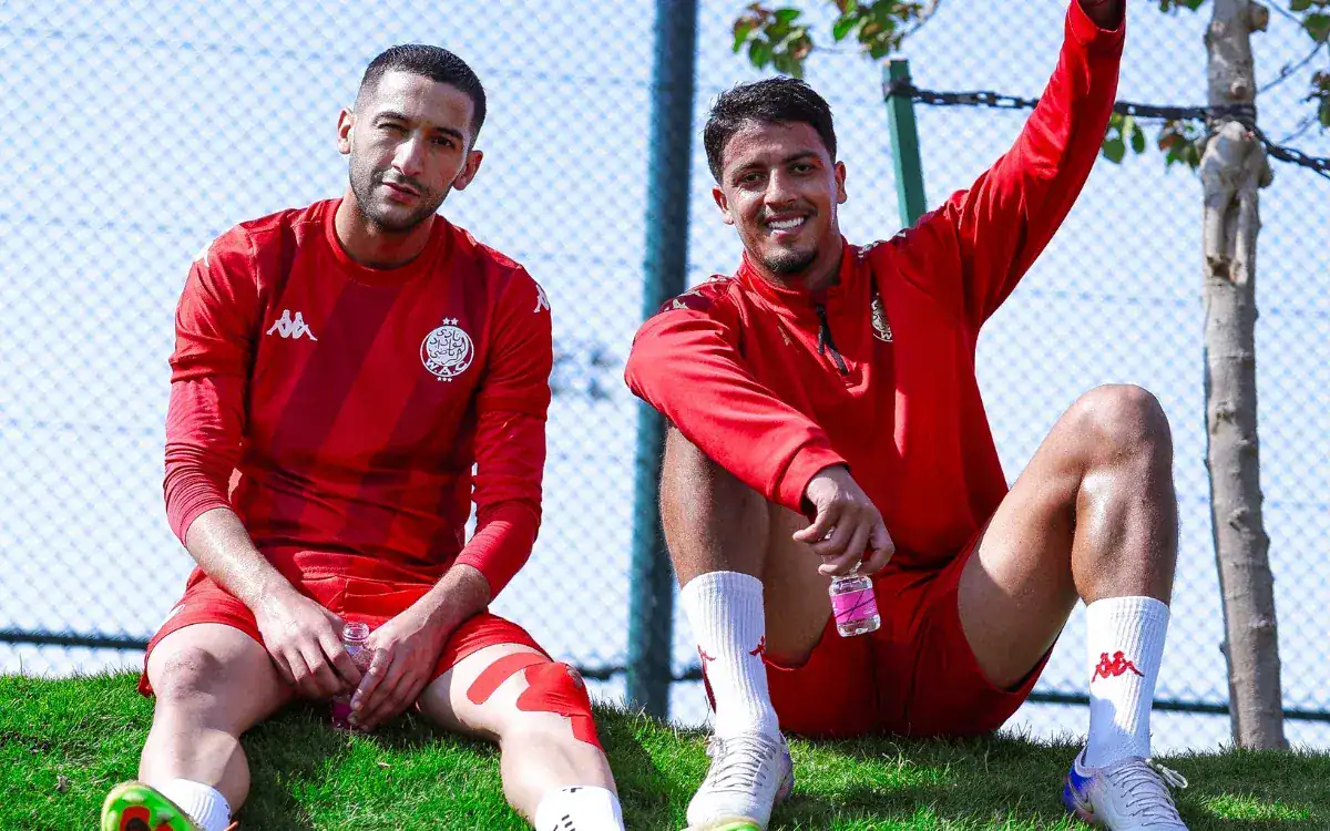 Hakim Ziyech et un coéquipier se détendent sur l'herbe, tous deux vêtus de tenues d'entraînement rouges, profitant d'une journée ensoleillée lors d'une séance d'entraînement de l'équipe marocaine.