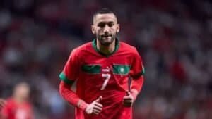 Hakim Ziyech, portant le maillot rouge du Maroc, numéro 7, faisant un signe de pouce levé pendant un match, illustrant les couleurs vibrantes et l'esprit de l'équipe.