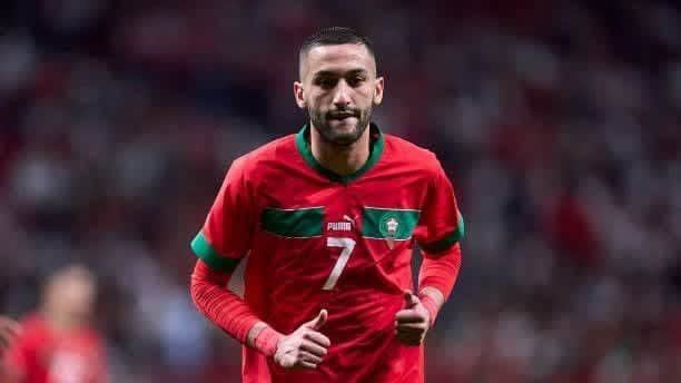 Hakim Ziyech, portant le maillot rouge du Maroc, numéro 7, faisant un signe de pouce levé pendant un match, illustrant les couleurs vibrantes et l'esprit de l'équipe.