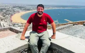 Smiling man in a red t-shirt sits on a stone ledge with a panoramic view of a sandy beach and blue sea in the background.