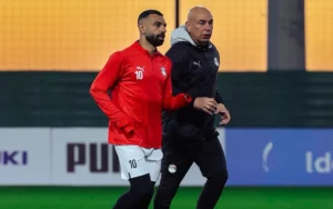 Hossam Hassan, portant un maillot d'entraînement rouge de l'Égypte, marche avec son entraîneur lors d'une séance d'entraînement de football, soulignant la camaraderie de l'équipe égyptienne.