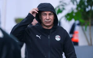 Hossam Hassan portant un sweat à capuche noir Puma, ajustant sa capuche, avec l'emblème de l'équipe nationale d'Égypte visible, sur fond flou.