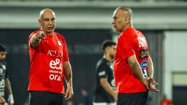 Two football coaches, both in red training kits, discussing strategies on the pitch, with players blurred in the background.