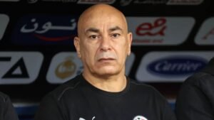 Hossam Hassan, entraîneur de l'équipe nationale égyptienne, fixant intensément le terrain depuis le banc pendant un match, avec les logos de l'équipe en arrière-plan.