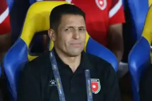 Hussein Ammouta, portant une chemise de coaching noire avec un emblème de la Jordanie, est assis pensivement dans un stade avec des sièges jaunes derrière lui.