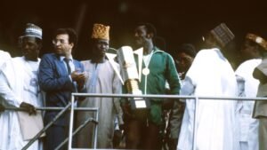 Célébrant avec un trophée, un joueur en survêtement vert se tient fièrement parmi des officiels en tenue traditionnelle lors d'un événement sportif majeur.