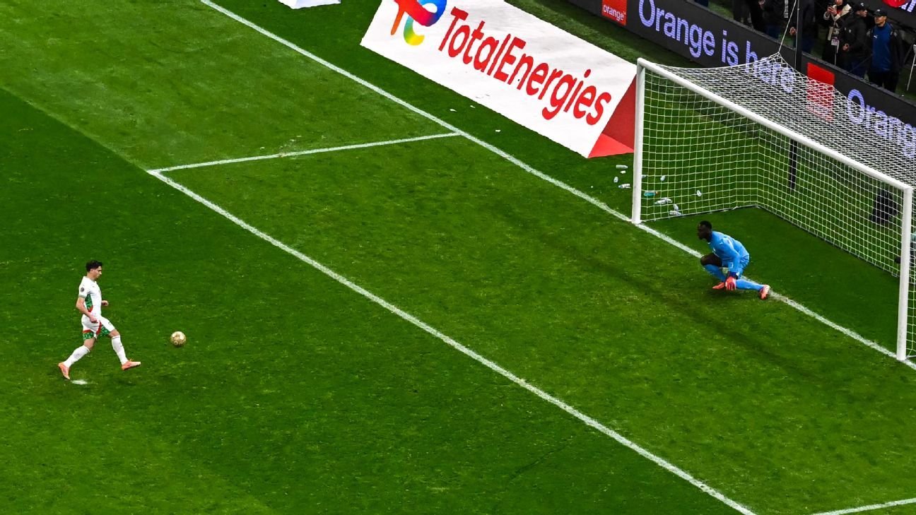 Moroccan player prepares to take a penalty kick with the goalkeeper crouching in anticipation, showcasing vibrant green pitch and advertising.