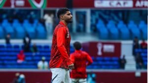 Ibrahim Adel in a red training kit, featuring black patterns, walking on the pitch with empty blue stadium seats in the background.