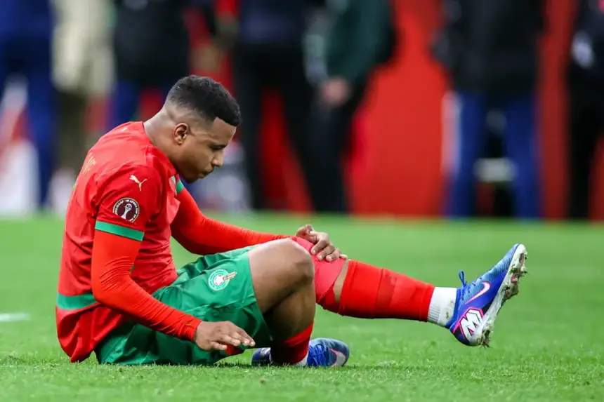 Moroccan player sits on the pitch, looking down at his leg in distress, wearing a red and green kit during a tense match moment.