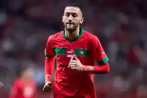 Hakim Ziyech portant le maillot rouge de l'équipe nationale marocaine, numéro 7, souriant et courant sur le terrain pendant un match, avec une foule animée en arrière-plan.