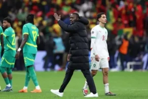 Moroccan and Senegalese players on the pitch, with a Senegalese coach gesturing. Intense match atmosphere with fans in the background.