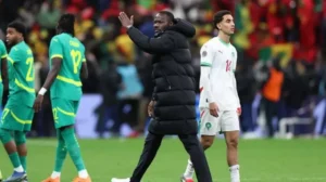 Moroccan coach gesturing on the sidelines as players walk past in a match against Senegal, with fans in the background.