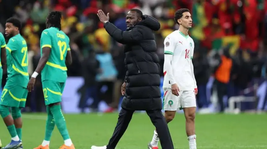 Moroccan coach gesturing on the sidelines as players walk past in a match against Senegal, with fans in the background.