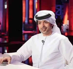 Smiling man in traditional Emirati attire sits at a desk with a smartphone, surrounded by a vibrant red and blue studio backdrop.