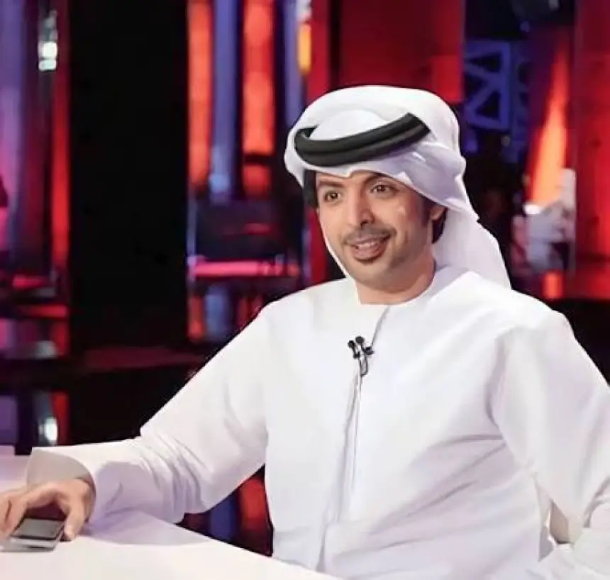 Smiling man in traditional Emirati attire sits at a desk with a smartphone, surrounded by a vibrant red and blue studio backdrop.