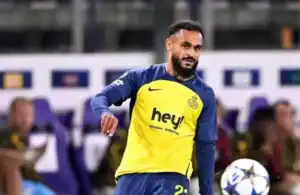 Joueur marocain en maillot jaune et bleu avec le logo 'hey!', contrôlant un ballon lors d'un match, avec des supporters visibles en arrière-plan.