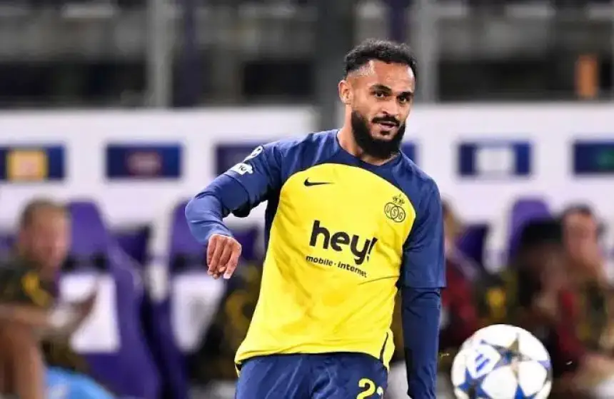Joueur marocain en maillot jaune et bleu avec le logo 'hey!', contrôlant un ballon lors d'un match, avec des supporters visibles en arrière-plan.