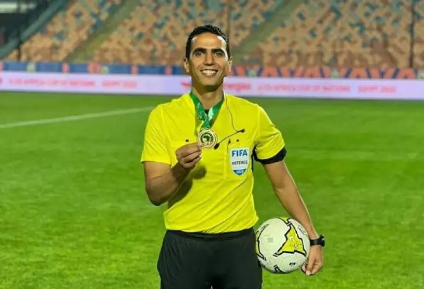 FIFA referee proudly holds a medal and a match ball, wearing a yellow uniform on a football pitch, celebrating a key achievement.