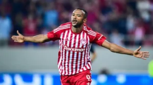 Moroccan striker celebrating in red and white striped Olympiacos kit, arms outstretched in a jubilant stadium atmosphere.