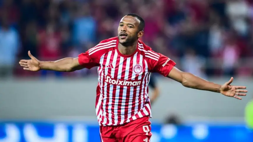 Moroccan striker celebrating in red and white striped Olympiacos kit, arms outstretched in a jubilant stadium atmosphere.