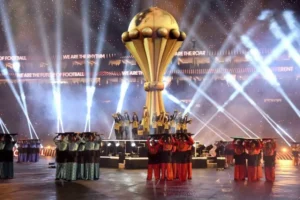Feestelijke scène van de AFCON-openingsceremonie met een grote trofee, artiesten in traditionele kleding en dramatische lichteffecten.