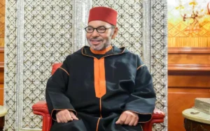 Koning Mohammed VI seated, wearing a traditional black robe with orange accents, in an ornate room with intricate wall décor.