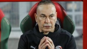 L'entraîneur de football marocain, plongé dans ses pensées en bord de terrain, porte un survêtement noir Puma, l'air concentré.