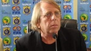 Een in gedachten verzonken coach, gezeten aan een vergadertafel, met een achtergrond van voetbalbondlogo's waaronder CAF en FIFA, besprekend inzichten over Marokkaans voetbal.