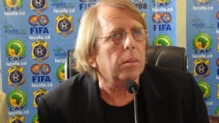 Een in gedachten verzonken coach, gezeten aan een vergadertafel, met een achtergrond van voetbalbondlogo's waaronder CAF en FIFA, besprekend inzichten over Marokkaans voetbal.