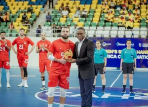 Un joueur marocain de handball portant le maillot rouge #9 reçoit un prix d'un officiel, avec ses coéquipiers et les arbitres en arrière-plan.