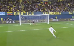 Kylian Mbappé bereidt zich voor op een penalty in een vol stadion, terwijl de doelman een duik maakt om te redden. Fel geel en blauw domineren de achtergrond.