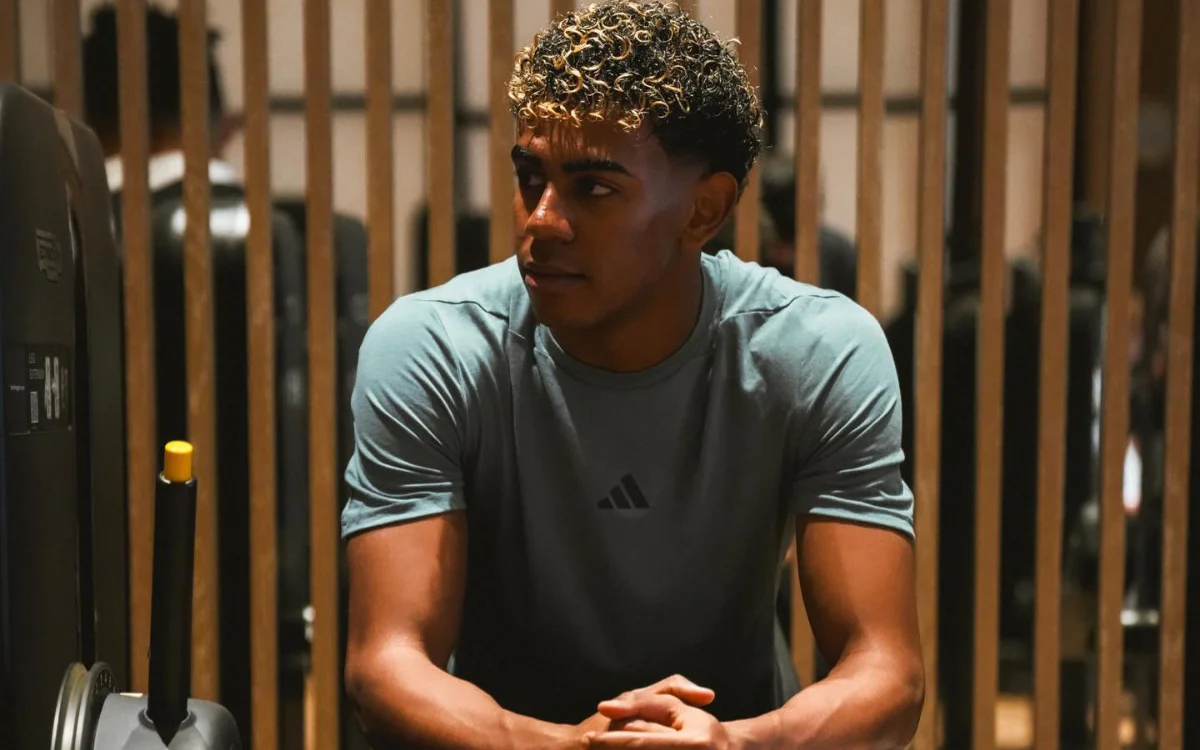 لامين يامال يجلس متفكراً في صالة رياضية، مرتدياً قميص أديداس فيروزي، يبرز ذراعيه المتناسقة على خلفية من الألواح الخشبية.