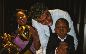 Lamine Yamal pose de manière ludique avec un enfant, tous deux souriants, tandis qu'une femme en tenue élégante tient un trophée, célébrant une réussite footballistique.