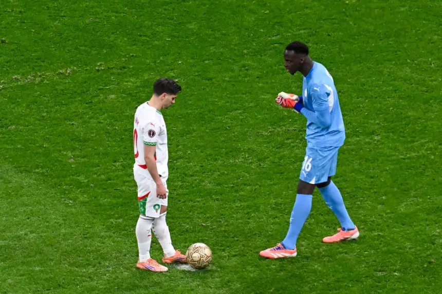 Un joueur marocain se tient au-dessus d'un ballon tandis que le gardien de but fait un geste, sur fond de pelouse d'un vert éclatant lors d'un match international.