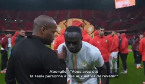 Sadio Mané talks with a coach before a match, surrounded by players in red jerseys, with a stadium backdrop showcasing Moroccan football pride.
