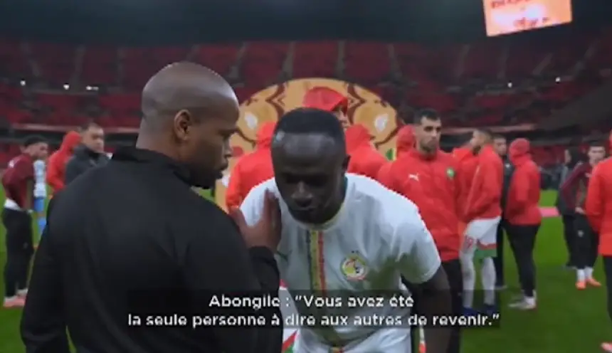 Sadio Mané talks with a coach before a match, surrounded by players in red jerseys, with a stadium backdrop showcasing Moroccan football pride.