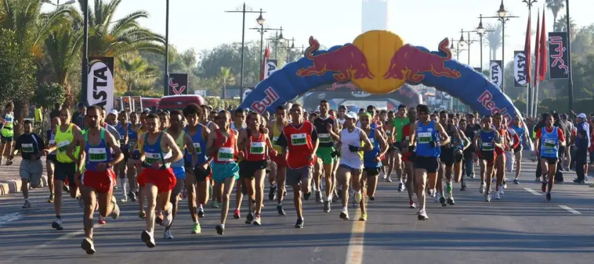 Runners in vibrant kits sprint towards a Red Bull arch at a Moroccan marathon, lined with palm trees and flags, in bright morning light.