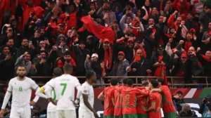 Des supporters marocains en rouge vif célèbrent tandis que les joueurs s'embrassent de joie après un but lors d'un match contre l'Algérie, reflétant la fierté nationale.