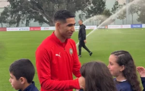 Achraf Hakimi, vêtu du maillot rouge du Maroc, interagit avec des enfants enthousiastes sur un terrain d'entraînement, avec des arroseurs en arrière-plan.