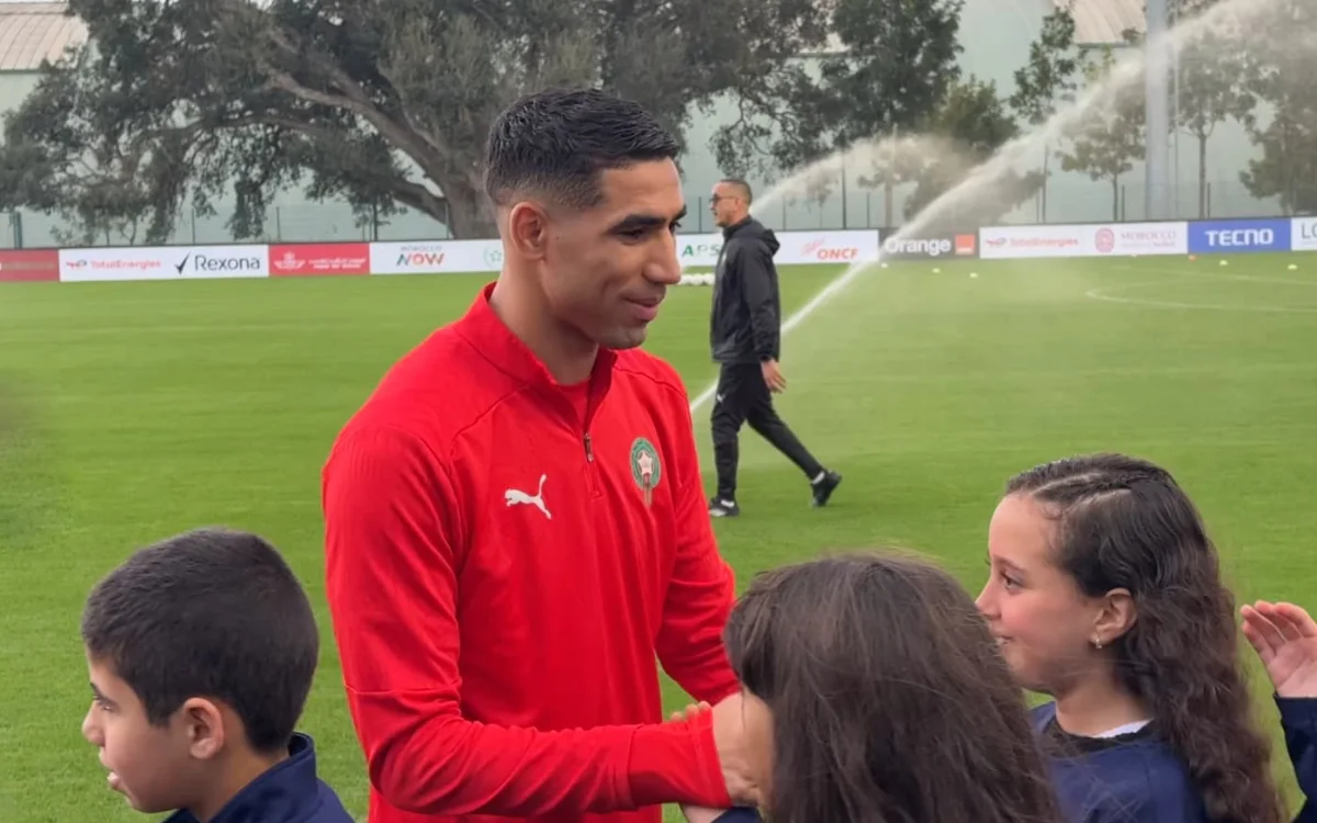 Achraf Hakimi, vêtu du maillot rouge du Maroc, interagit avec des enfants enthousiastes sur un terrain d'entraînement, avec des arroseurs en arrière-plan.