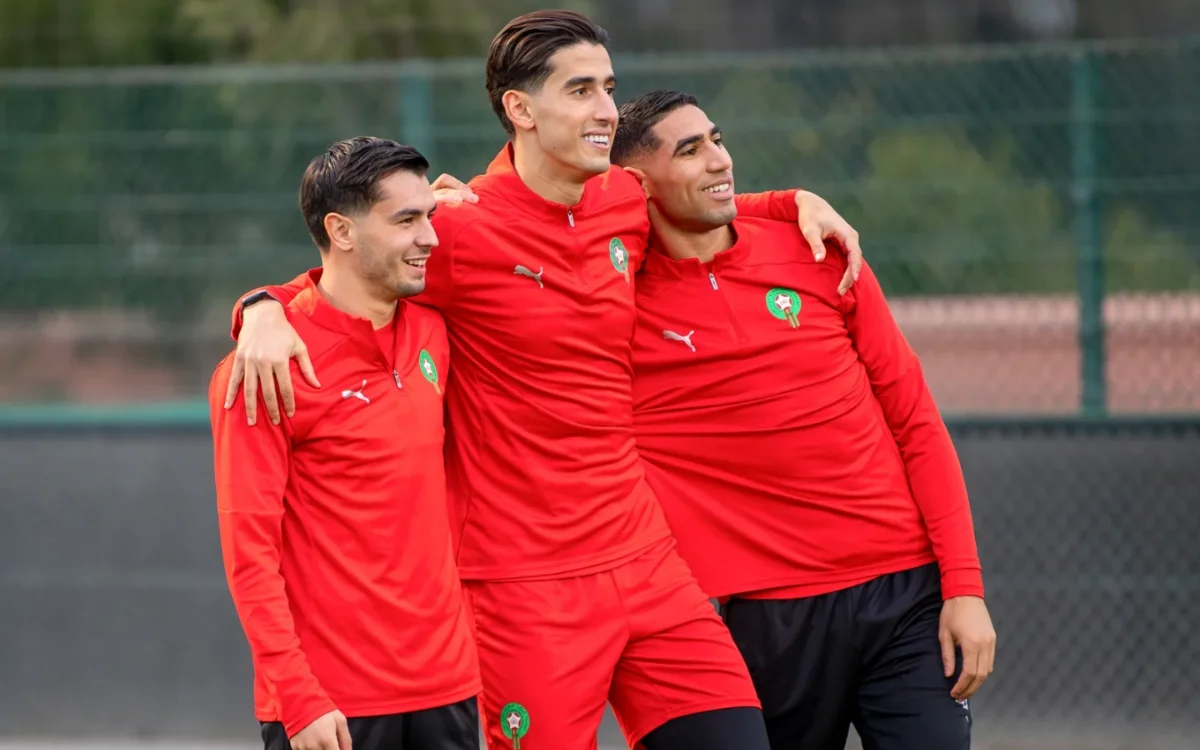 Des footballeurs marocains s'embrassent pendant l'entraînement, vêtus de tenues rouges Puma arborant les emblèmes nationaux, illustrant l'esprit d'équipe avant un match.