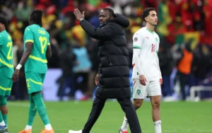 Morocco's coach gestures as Moroccan player (14) walks past; Senegal players in vibrant green uniforms amid a spirited stadium crowd.