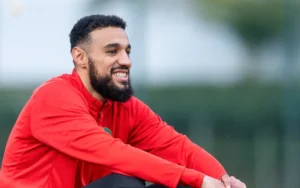 Moroccan footballer smiling in red training kit, seated on grass, showcasing camaraderie and focus during a training session.