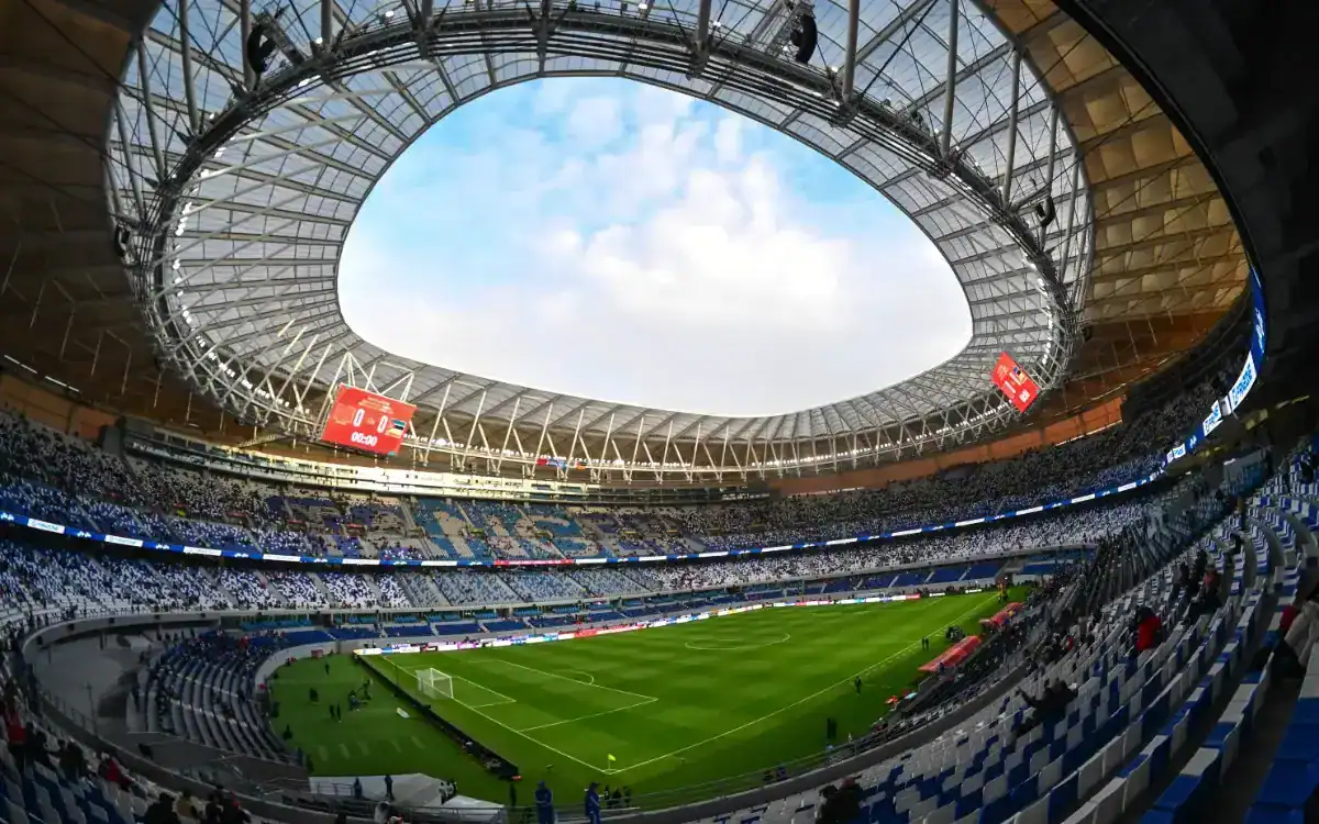 Intérieur moderne d'un stade avec un terrain vert, des gradins bleus et blancs, et un ciel clair en plafond, des écrans numériques affichant les informations du match.