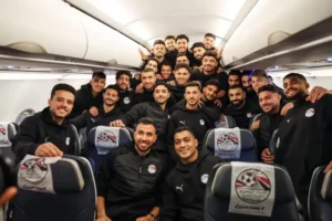Egyptian national football team players pose for a group photo on an airplane, wearing black tracksuits, smiling and celebrating together.