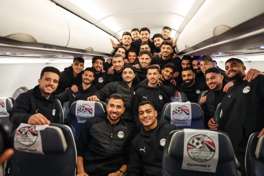 Egyptian national football team players pose for a group photo on an airplane, wearing black tracksuits, smiling and celebrating together.
