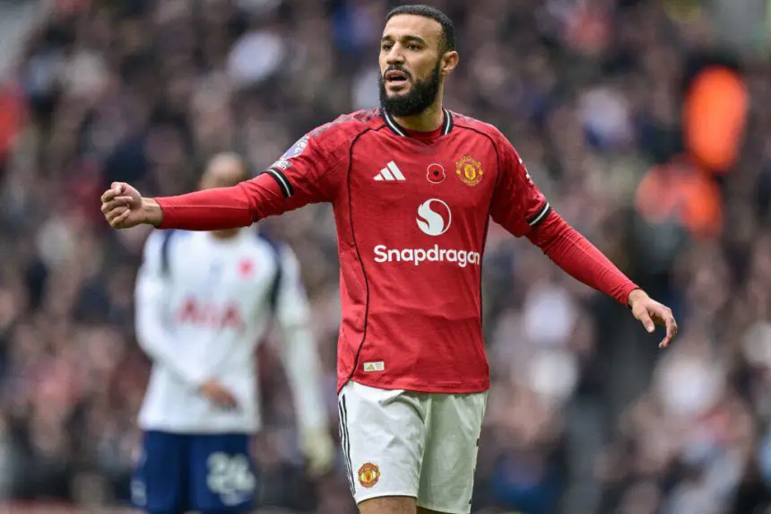 Le joueur marocain Hakim Ziyech dans le maillot rouge de Manchester United gesticule pendant un match, avec des fans et des joueurs adverses en arrière-plan.