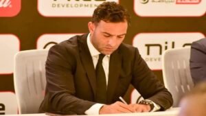 Mido, wearing a formal black suit, signs a document at a press conference, focused amidst an official backdrop with branding.